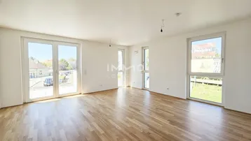 Heller, leerer Wohnraum mit großen Fenstern, Holzboden und Zugang zu einem Außenbereich.