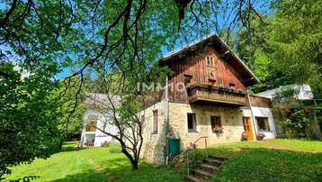 Traditionelles Haus mit Holzbalkon und Steinfassade, umgeben von üppigem Grün und Bäumen.