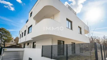 Modernes weißes Gebäude mit geschwungenen Balkonen und Tiefgaragenzufahrt unter blauem Himmel.