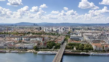 Weitläufige Luftaufnahme einer Stadt mit Fluss, Brücke und Bergen im Hintergrund unter blauem Himmel.