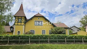 Historisches Gebäude mit markantem Turm, gelber Fassade und gepflegter Hecke zur Straße hin.