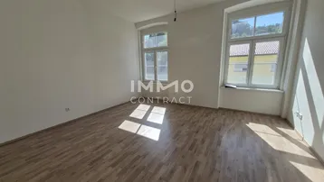 Helles, leeres Zimmer mit Holzboden und großen Fenstern, das viel Tageslicht hereinlässt.
