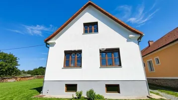 Weißes Einfamilienhaus mit braunen Fensterrahmen und rotem Ziegeldach auf einem grünen Grundstück.