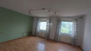 Helles Zimmer mit Parkettboden, grüner Akzentwand und drei Fenstern, die viel Tageslicht hereinlassen.