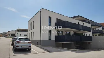 Modernes Mehrfamilienhaus mit weißer Fassade, dunklen Balkonen und Tiefgaragenzufahrt in sonniger Lage.