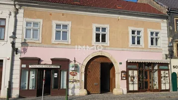 Historisches Gebäude mit beiger und rosa Fassade, Holztor und Geschäften im Erdgeschoss an einer Kopfsteinpflasterstraße.