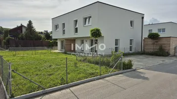 Modernes weißes Gebäude mit schlichter Architektur, umgeben von einer Rasenfläche und einem Parkplatz.