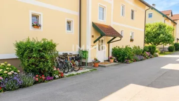 Gelbes Mehrfamilienhaus mit blühenden Blumenbeeten und Fahrrädern vor dem Eingang.