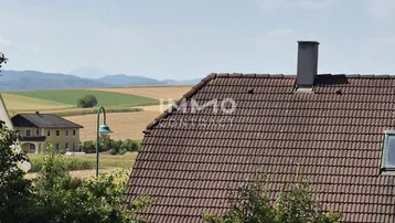 Weitläufiger Blick über Felder und Dächer mit Bergen im Hintergrund.