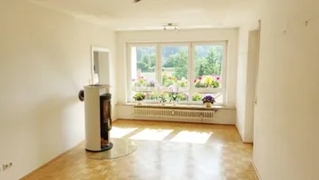 Helles Wohnzimmer mit Parkettboden, Kaminofen und großen Fenstern mit Blick ins Grüne.