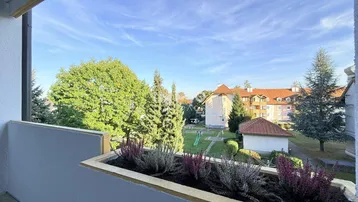 Blick vom Balkon auf eine grüne Wohnanlage mit Bäumen und Spielplatz unter blauem Himmel.