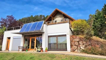 Modernes Haus mit Solarpaneelen auf dem Dach und Balkon in ländlicher Umgebung.