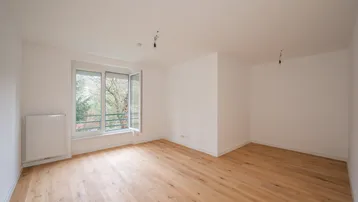 Helles, leeres Zimmer mit Parkettboden und großem Fenster, das viel Tageslicht hereinlässt.