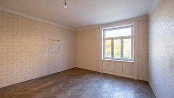 Leeres Zimmer mit Fischgrätparkett, gemusterter Tapete und einem großen Fenster, das viel Licht hereinlässt.