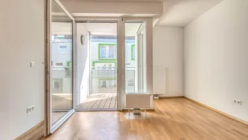 Helles Zimmer mit Parkettboden und großen Glasfronten, die Zugang zu einem Balkon bieten.