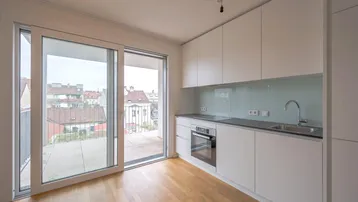 Moderne Einbauküche mit weißen Schränken, dunkler Arbeitsplatte und Zugang zu einem großen Balkon.