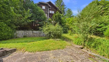Gartenbereich mit Rasenfläche, Steinmauer und Blick auf das Haus durch die Bäume.