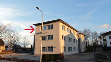 Modernes Bürogebäude mit heller Fassade und Parkplätzen im Freien unter blauem Himmel, gut sichtbar von der Straße.