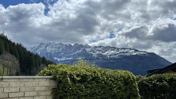 Panoramablick auf schneebedeckte Berge und grüne Wälder unter einem bewölkten Himmel.