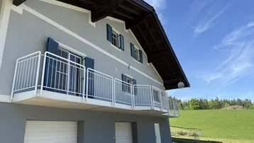 Traditionelles Haus mit Balkon und Garagen im Erdgeschoss, umgeben von grüner Landschaft.