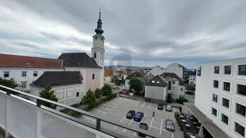 Blick von einem Balkon auf eine Stadtansicht mit Kirche, Parkplatz und umliegenden Gebäuden unter bewölktem Himmel.
