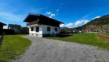 Einfamilienhaus mit Schotterzufahrt und Rasenfläche vor alpiner Bergkulisse.