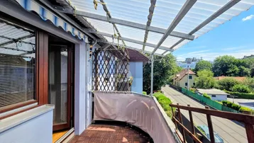 Überdachter Balkon mit Holzboden und Blick auf die Straße und umliegende Bäume.