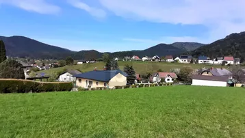Panoramablick auf eine ländliche Siedlung mit Häusern, umgeben von grünen Wiesen und Bergen unter blauem Himmel.