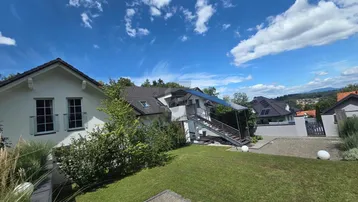 Modernes Haus mit gepflegtem Garten, Photovoltaikanlage und weitem Blick unter blauem Himmel.