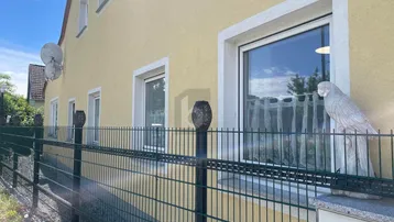 Seitenansicht eines gelben Hauses mit weißen Fenstern und einem grünen Metallzaun im Vordergrund.