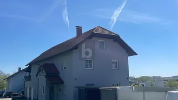 Graues Einfamilienhaus mit Satteldach und kleinem Vordach unter blauem Himmel.