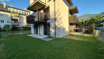 Modernes Gebäude mit Balkonen und einem gepflegten Rasenbereich, umgeben von grüner Landschaft und Bergen.