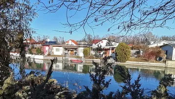 Blick über einen ruhigen Kanal auf gegenüberliegende Häuser und üppige Vegetation unter blauem Himmel.