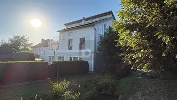 Das Haus präsentiert sich mit einer modernen Fassade, umgeben von einem gepflegten Garten und einem kleinen Bachlauf.