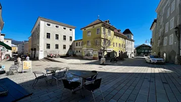 Ein sonniger Stadtplatz mit historischen Gebäuden und Außengastronomie, umgeben von Bergen.