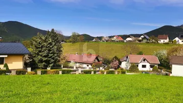 Panoramablick auf eine ländliche Siedlung mit Häusern und grünen Hügeln im Hintergrund unter blauem Himmel.