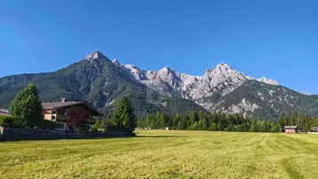 Ein Haus in einer weiten grünen Wiese mit majestätischen Bergen im Hintergrund unter blauem Himmel.