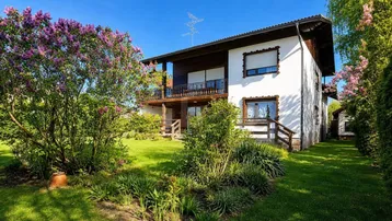 Weißes Einfamilienhaus mit gepflegtem Garten und blühenden Fliederbüschen unter blauem Himmel.