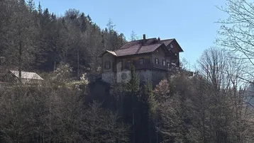 Traditionelles Holzhaus auf einem Hügel, umgeben von Wald und Natur unter blauem Himmel.