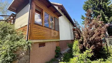 Seitenansicht des Hauses mit Holzverkleidung, Fenstern und üppiger grüner Umgebung unter blauem Himmel.