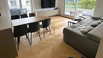 Helles Wohn- und Esszimmer mit Fischgrätparkett, großen Fenstern und Zugang zum Balkon.