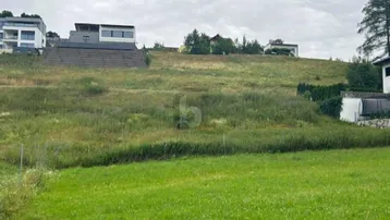 Grünes Baugrundstück an einem Hang mit modernen Häusern im Hintergrund unter bewölktem Himmel.
