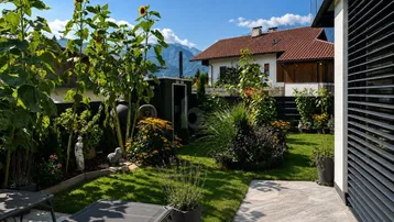 Gepflegter Garten mit Rasenfläche, Sonnenblumen und einer Terrasse mit Bergblick.