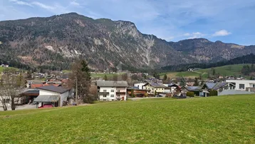Grüne Wiesen im Vordergrund mit einem Dorf und majestätischen Bergen im Hintergrund unter blauem Himmel.
