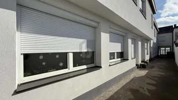 Fassade eines Wohngebäudes mit geschlossenen Rollläden und einem schmalen Zugangsweg.