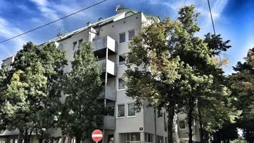 Modernes Mehrfamilienhaus mit Balkonen und großen Fenstern, umgeben von Bäumen an einer städtischen Straße.