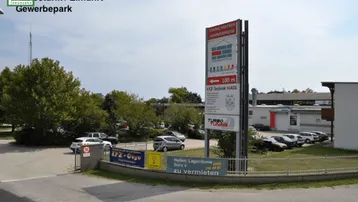 Einfahrt zu einem Gewerbepark mit großem Werbeschild, geparkten Autos und Bäumen unter blauem Himmel.