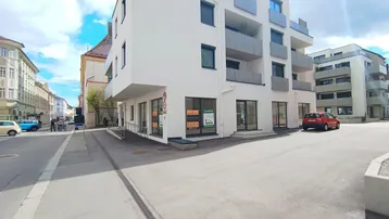 Modernes Eckgebäude mit großen Schaufenstern im Erdgeschoss und Balkonen in den oberen Etagen.