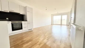 Heller offener Wohnbereich mit integrierter Küche, Parkettboden und Zugang zum Balkon.