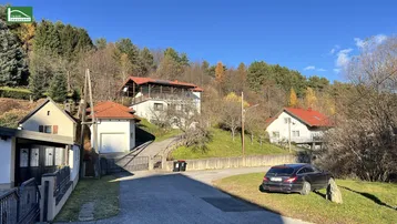 Blick auf mehrere Häuser in einer ländlichen Umgebung mit bewaldeten Hügeln unter blauem Himmel.